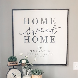 May include: A wooden sign with the text "Home sweet Home the McEvoy's Established 2005" in black lettering. The sign is hanging on a gray wall above a wooden cabinet with drawers. The cabinet is decorated with plants and a vintage scale.