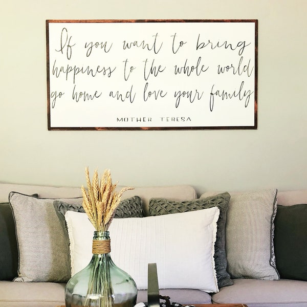 May include: A wooden sign with the quote "If you want to bring happiness to the whole world go home and love your family" by Mother Teresa. The sign is hanging on a light green wall above a couch with pillows and a coffee table with a decorative tray.