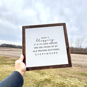May include: A square wooden sign with a white background and a dark brown frame. The sign reads "WHAT A blessing IT IS TO LOOK AROUND AND SEE PIECES OF MY OLD PRAYERS SCATTERED EVERYWHERE" in black lettering.