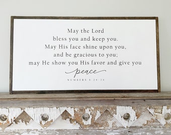 May include: A white wooden sign with a brown frame, featuring a religious quote: "May the Lord bless you and keep you. May His face shine upon you, and be gracious to you; may He show you His favor and give you peace." The quote is attributed to Numbers 6:24-26.
