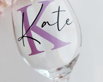 Puede incluir: Una copa de vino de vidrio transparente con una pegatina de vinilo morado que dice "Kate" con una gran "K" morada en letra cursiva.
