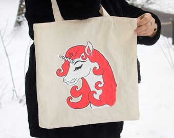 Puede incluir: Una bolsa de tela blanca con una ilustración de unicornio rojo y blanco. El unicornio tiene una crin y una cola rojas y un cuerpo blanco. El unicornio está mirando hacia la izquierda.