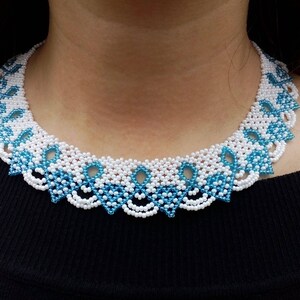 Op de afbeelding: Een witte en blauwe kralenketting met een delicaat, ingewikkeld ontwerp. De ketting is gemaakt van kleine, ronde kralen die aan elkaar geregen zijn in een patroon van bloemen en bladeren.