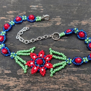 May include: A beaded bracelet with a red flower charm. The bracelet is made with green, blue, and red beads. The flower charm has a red center and white and blue petals.