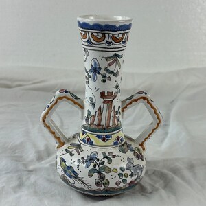 May include: A white ceramic vase with a bulbous base and two handles. The vase is decorated with hand-painted floral and architectural designs in blue, green, yellow, and brown. The rim is trimmed with a blue and orange pattern.