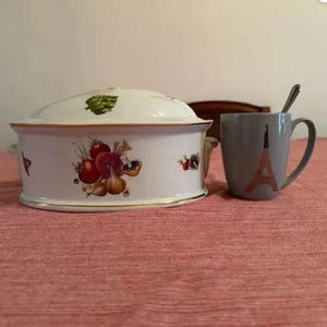 Może przedstawiać: Biała ceramiczna salaterka z pokrywką, ozdobiona kolorowymi ilustracjami warzyw. Obok naczynia stoi szary kubek z wizerunkiem Wieży Eiffla i łyżeczką w środku. Naczynie ma złote obramowanie.