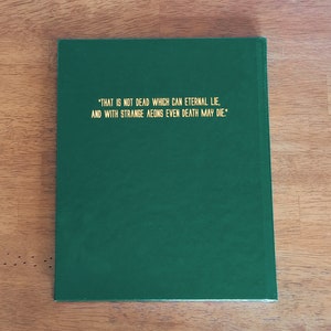 May include: A dark green hardcover book with gold lettering on the front cover. The text reads: "THAT IS NOT DEAD WHICH CAN ETERNAL LIE, AND WITH STRANGE AEONS EVEN DEATH MAY DIE."