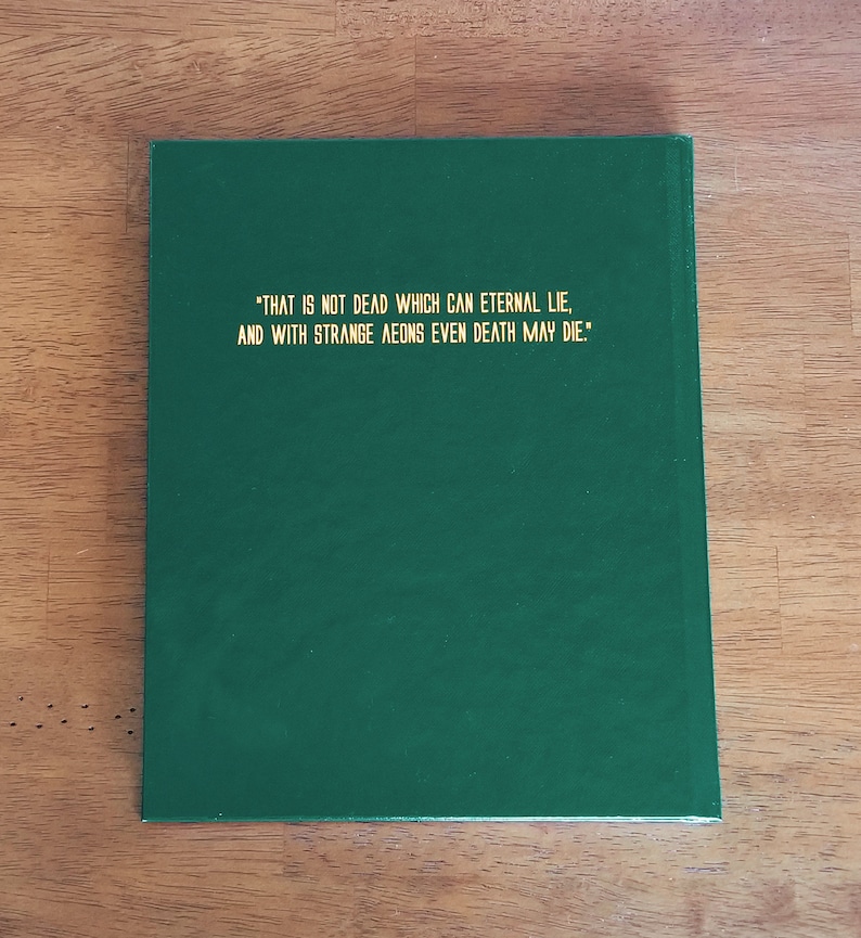 May include: A dark green hardcover book with gold lettering on the front cover. The text reads: "THAT IS NOT DEAD WHICH CAN ETERNAL LIE, AND WITH STRANGE AEONS EVEN DEATH MAY DIE."