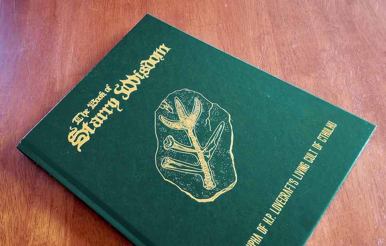 May include: A green hardcover book with gold lettering titled "The Book of Starry Wisdom" and a gold illustration of a fossil on the front cover. The bottom of the cover reads "Cthulhu of H.P. Lovecraft's Living Cult of Cthulhu."