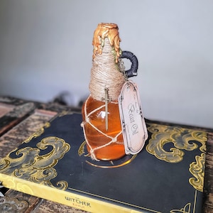 Może przedstawiać: Ozdobna szklana butelka wypełniona bursztynowym płynem, owinięta sznurkiem i uszczelniona złotym woskiem. Dołączony jest mały urok miecza i etykieta z napisem "Beast Oil". Butelka stoi na książce ze złotymi akcentami.
