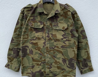 May include: A green, brown, and black camouflage long-sleeved shirt with a button-up front and two flap pockets. The shirt is hanging on a hanger.