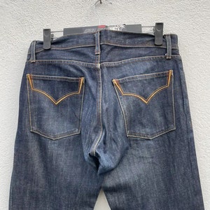 Op de afbeelding: Een paar donkerblauwe spijkerbroeken met gele stiksels op de achterzakken. De jeans heeft een klassieke vijf-zak stijl.