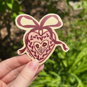 May include: A heart-shaped sticker with a bow and ribbon design. The sticker features a burgundy heart with floral and leaf details, and a cream-colored background. The sticker is held by a hand against a blurred green background.