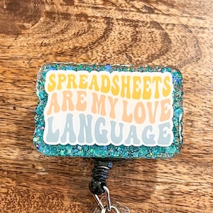 May include: A teal blue, glittery badge holder with the text "Spreadsheets are my love language" in a retro font. The badge holder has a black retractable clip and a clear plastic badge holder.