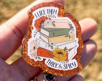 Può includere: Un portabadge di glitter marrone e arancione con una pila di libri e fiori. Il testo sul portabadge recita "I like them thick & spicy".