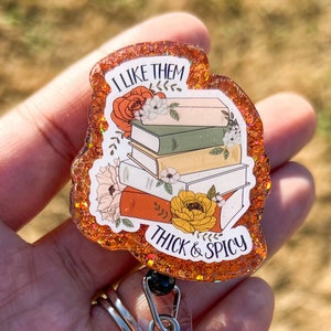 May include: A brown and orange glitter badge holder with a stack of books and flowers. The text on the badge holder reads "I like them thick & spicy".