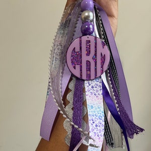 Puede incluir: Llavero de borla morado y plateado con un gran charm circular de purpurina morado con el monograma "CRM" en blanco.