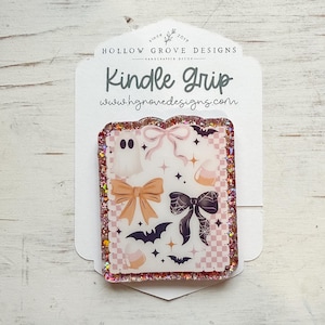 May include: A rectangular Kindle grip with a glittery pink border. The grip features a Halloween-themed design with a ghost, bows, bats, and candy corn on a pink and white checkered background. The text "Kindle Grip" and the website address are visible.