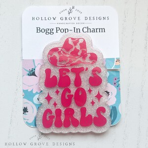Puede incluir: Un charm brillante rosa y blanco con el texto "LET'S GO GIRLS" y un diseño de sombrero de vaquera rosa. El charm está unido a una tarjeta blanca con el texto "HOLLOW GROVE DESIGNS" y "Bogg Pop-In Charm".