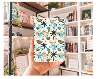 Könnte beinhalten: Eine gemusterte Hülle mit tropischem Design. Sie zeigt Palmen, Margarita-Gläser und Limettenscheiben auf weißem Hintergrund mit blauen, grünen und gelben Akzenten. Die Hülle wird in einer Hand gehalten.