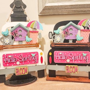 May include: Two wooden truck-shaped spring decorations with a pink and white polka dot background. The trucks have a birdhouse, a watering can, a butterfly, and a rain boot on the back. The trucks say "Hello Spring" and "Bloom".