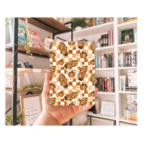 Könnte beinhalten: Eine Tablet-Hülle mit einem sich wiederholenden Muster aus Kürbissen, Kaffeetassen und Sternen auf einem karierten orange-weißen Hintergrund. Die Hülle wird in einer Hand gehalten, mit einem Bücherregal im Hintergrund.