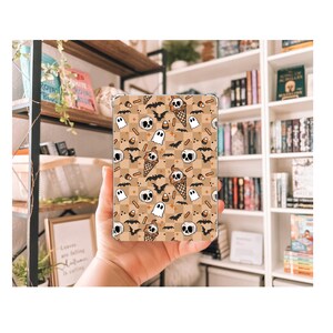 May include: A clear case featuring a Halloween-themed pattern. The design includes skulls, bats, ghosts, and ice cream cones with skull faces, set against a tan and brown checkered background. The case is held in a hand.
