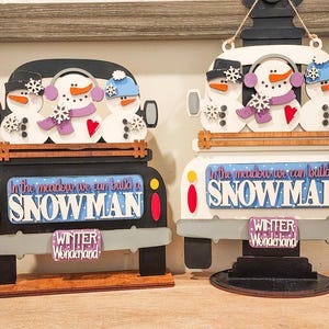 以下が含まれることがあります： 雪だるまが描かれた木製のトラックのオーナメント2つ。"In the meadow we can build a snowman"と"Winter Wonderland"の文字が書かれている。