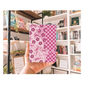 May include: A clear tablet case with a pink floral and bow pattern on one side and a pink checkered pattern on the other. The case is held in a hand, with a bookshelf in the background.