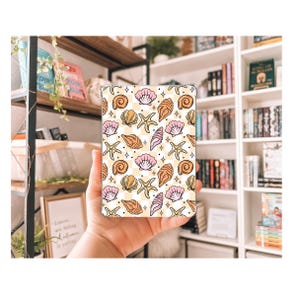 以下が含まれることがあります： 長方形の携帯電話ケース。桃色、ピンク、タンの色合いの貝殻、ヒトデ、巻貝の繰り返し模様が、チェック柄の背景に配置されています。デザインには小さな星型のアクセントが含まれています。
