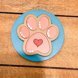 May include: A light blue wooden circle with a pink paw print in the center. The paw print has a small red heart in the middle.