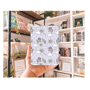 Puede incluir: Una funda de teléfono de color morado y blanco con un patrón floral de margaritas blancas, flores moradas y hojas verdes. El fondo es de color morado claro con nubes blancas.