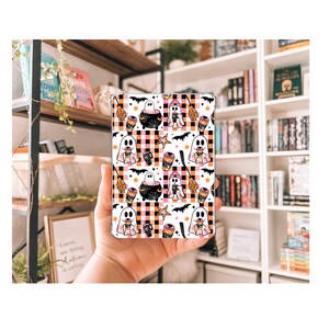 Könnte beinhalten: Eine gemusterte Handyhülle mit Halloween-Motiv. Das Design zeigt Geister, Fledermäuse, Sterne, Kessel und Kaffeetassen auf einem schwarz-weiß karierten Hintergrund. Die Hülle wird in der Hand einer Person gehalten.