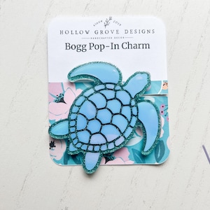 Könnte beinhalten: Ein hellblaues, schildkrötenförmiges Bogg Pop-In Charm mit glitzernder Kontur, präsentiert auf einer Karte mit dem Text "HOLLOW GROVE DESIGNS" und "Bogg Pop-In Charm". Der Hintergrund zeigt ein Blumenmuster.