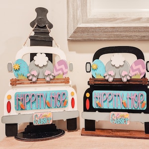 Puede incluir: Dos decoraciones de camión de Pascua de madera, una blanca y otra negra, con una cola de conejo que sobresale de la parte trasera. Ambos camiones tienen huevos de Pascua y las palabras "Hippity Hop" y "Easter" en ellos.