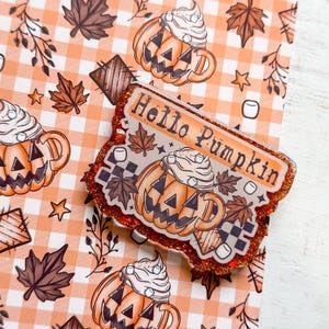 May include: An orange and brown "Hello Pumpkin" badge with glitter edges, featuring a pumpkin-shaped coffee cup design with whipped cream, leaves, and marshmallows. The background is a fall-themed pattern with pumpkins, leaves, and stars.