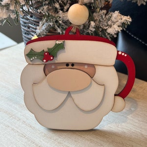 May include: Wooden Christmas ornament shaped like Santa's face with a red hat, white beard, and a brown nose. The ornament has a red and white mug handle.