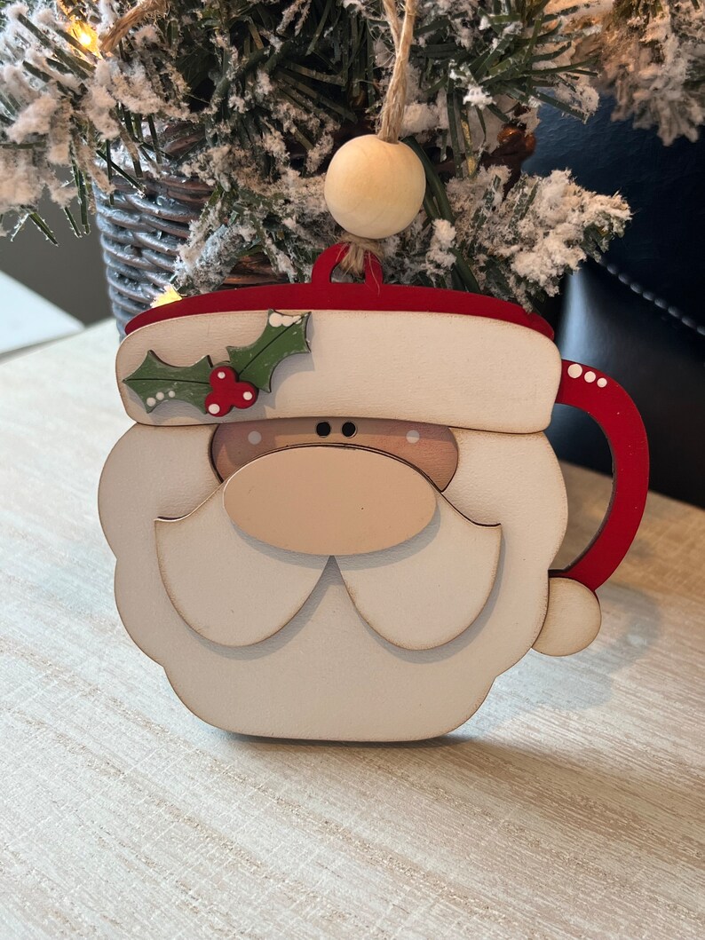 May include: Wooden Christmas ornament shaped like Santa's face with a red hat, white beard, and a brown nose. The ornament has a red and white mug handle.
