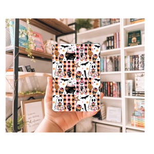 May include: A rectangular phone case with a Halloween pattern. The design features cartoon ghosts, cauldrons, coffee cups, bats, and stars on a black, white, and orange checkered background. The case is held in a person's hand.