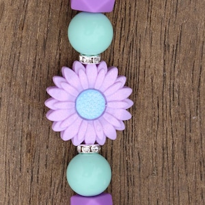 Puede incluir: Un llavero de cuentas de silicona de color morado y verde con un dije de flor y un cierre plateado.