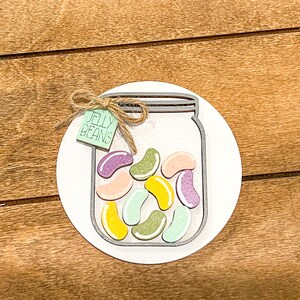 Puede incluir: Un círculo de madera blanco con un tarro de albañil pintado de gris lleno de caramelos de gelatina de colores. El tarro tiene una etiqueta verde que dice "Jelly Beans" atada con un cordel.