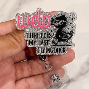Puede incluir: Un portatarjetas con un pato de dibujos animados en blanco y negro con gafas de sol. El portatarjetas tiene el texto "Welp! There goes my last flying duck" en rosa y negro. El portatarjetas está sujeto a un cordón retráctil.