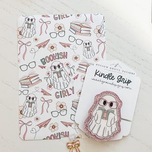 May include: A white and pink patterned book cover with the words "Bookish" and "Girly" and a Kindle Grip featuring a ghost reading a book. The Kindle Grip has a pink glitter border and the text "Kindle Grip". A gold bow charm is also present.