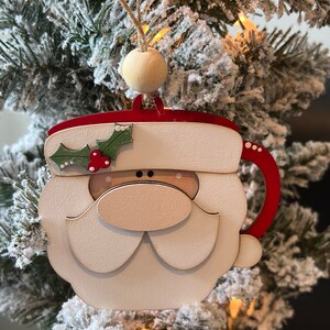 May include: A wooden Christmas bauble shaped like a Santa Claus mug. The mug is red and white with a white beard and a red nose. The ornament has a holly sprig with red berries on the side.
