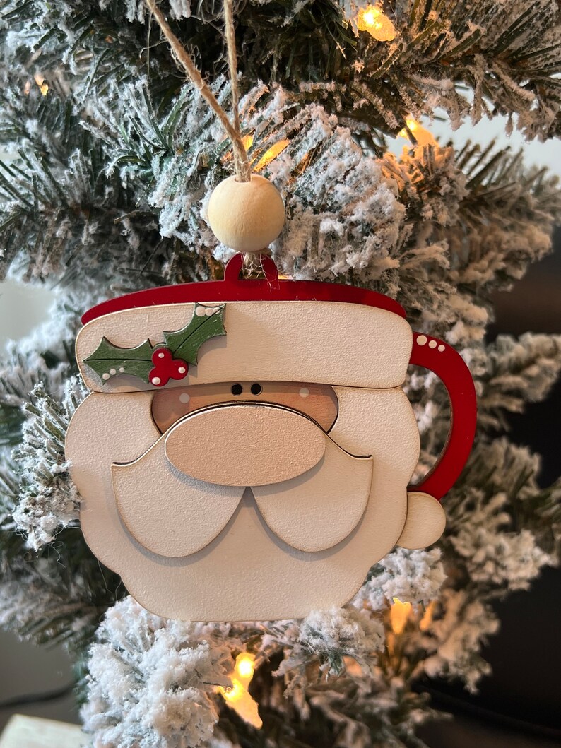 May include: A wooden Christmas bauble shaped like a Santa Claus mug. The mug is red and white with a white beard and a red nose. The ornament has a holly sprig with red berries on the side.
