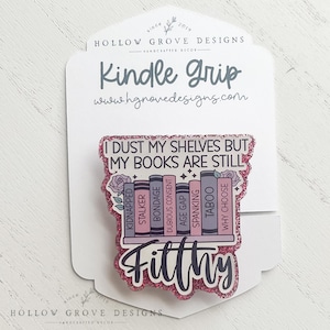 May include: A pink glitter Kindle Grip with the text "Kindle Grip" and "I DUST MY SHELVES BUT MY BOOKS ARE STILL Filthy". The design features a bookshelf with book titles like "Kidnapped" and "Taboo". The brand name "Hollow Grove Designs" is also visible.