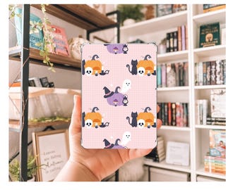 May include: A pink tablet case featuring a Halloween-themed pattern. The design includes orange pumpkins, black cats, white ghosts, and purple witch hats. The background has a grid pattern.