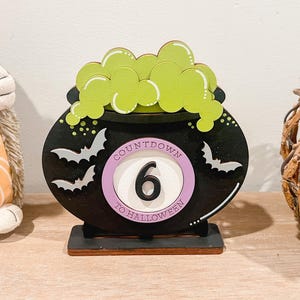 May include: A wooden Halloween countdown decoration shaped like a cauldron. The black cauldron has green bubbling potion, white bats, and a pink circle with the number 6 and the text "Countdown to Halloween".