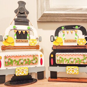 May include: Two wooden truck signs with a lemonade theme. The trucks are painted white and black with a pink and white lemonade sign on the back. Both trucks have a sign that says "Fresh Squeezed" and a price tag that says "25c".