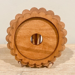 以下が含まれることがあります： 中央に円形の切り抜きがあり、縁が波打っている木製コースター。コースターは薄い茶色の木でできており、小さな木製スタンドの上に置かれています。スタンドには「Hollow Grove Designs」という文字が印刷されています。